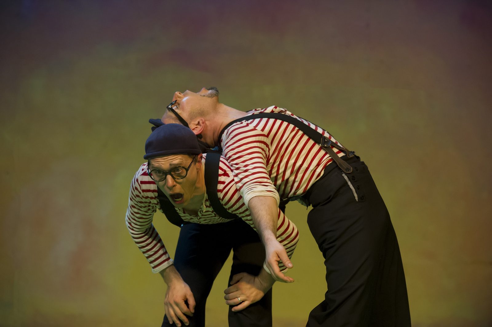 One performer carried in a deep backbend across the other's shoulders against an ochre painted backdrop